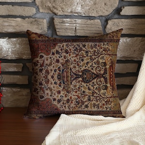 Puede incluir: Cojín decorativo cuadrado con un patrón detallado y ornamentado en tonos beige, marrón y rojo. El cojín está colocado sobre una superficie de madera frente a una pared de piedra y una manta crema.