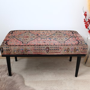 May include: A black wood bench with a pink and beige patterned rug top. The rug has a geometric design with a central diamond shape.