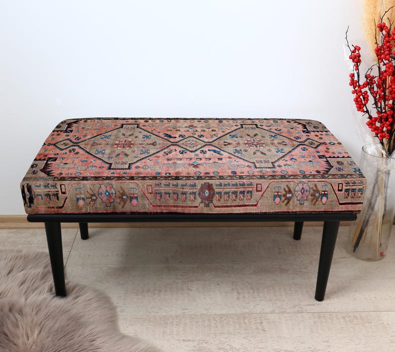 May include: A black wood bench with a pink and beige patterned rug top. The rug has a geometric design with a central diamond shape.