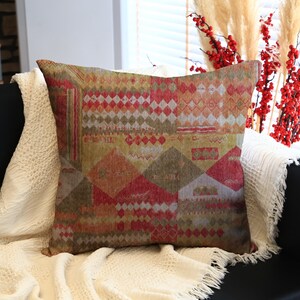 May include: A decorative throw pillow with a geometric pattern in shades of red, green, and beige. The pillow is square and rests on a black sofa, with a white blanket draped over it. Decorative plants are in the background.