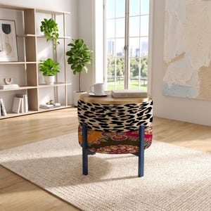 May include: A round ottoman with a two-tiered design featuring a leopard print top section and a colorful floral patterned bottom section. The piece is supported by four dark blue legs and topped with a flat, circular wooden surface.