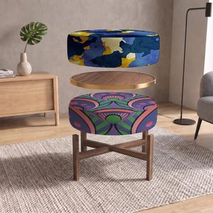May include: A round ottoman with a wooden base and a split design. The top section features a blue, yellow, and white camouflage pattern, while the bottom section displays a geometric print in shades of pink, green, and blue, separated by a wooden tray.