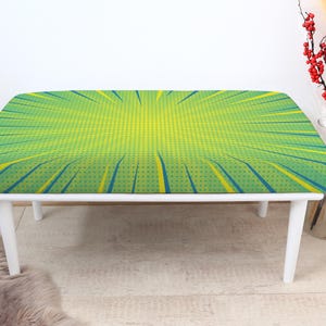 May include: A white-legged bench with a comic book-style design. The bench top features a green and yellow burst pattern with blue accents. The bench is rectangular with rounded corners and is supported by four white legs.