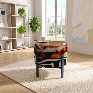 May include: A round ottoman with a patterned fabric cover featuring abstract shapes in orange, red, black, and cream. The piece has a flat, light-colored wooden top and is supported by four dark blue legs, functioning as a small side table.