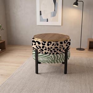 May include: A round ottoman with a wooden top and a gold metal rim. The upper section features a tan and black leopard print fabric, while the lower section has a green and white zebra print fabric. It stands on four black metal legs.