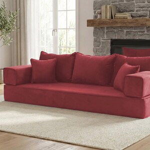 May include: A low-profile, red modular sofa with several matching throw pillows. The sofa is placed on a cream-colored rug in a living room setting. The room also features a stone fireplace, a wooden side table, and a potted olive tree.
