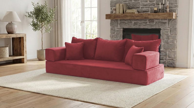 May include: A low-profile, red modular sofa with several matching throw pillows. The sofa is placed on a cream-colored rug in a living room setting. The room also features a stone fireplace, a wooden side table, and a potted olive tree.