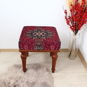 May include: A small, square ottoman with a burgundy patterned top and turned wooden legs. The patterned fabric features a central medallion design in shades of black, beige, and teal. The legs are a warm brown color.