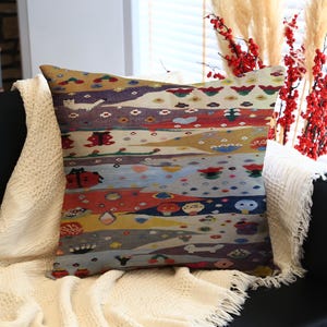 May include: Square decorative pillow with a colorful, abstract design. The pillow features horizontal bands of various colors, including red, blue, yellow, and gray, with whimsical patterns and shapes. It rests on a black couch with a white blanket.