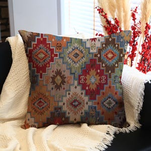 Könnte beinhalten: Ein quadratisches Zierkissen mit einem geometrischen Muster in Rot-, Orange-, Blau- und Brauntönen. Das Kissen liegt auf einer weißen Decke, die auf einem schwarzen Sofa platziert ist. Dekorative Zweige sind im Hintergrund.
