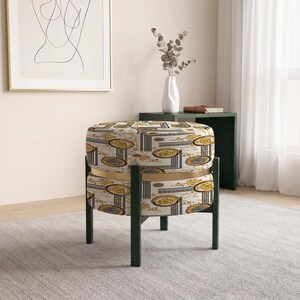 May include: A round ottoman featuring a cream-colored fabric with a repeating gold and black ornate pattern. The seat is divided by a thin gold metal band and is supported by four dark green legs that extend upward to frame the sides of the cushion.