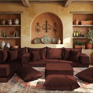 May include: A living room scene featuring a modular sectional sofa in a deep burgundy color. The room has a rustic, Mediterranean style with terracotta walls, wooden beams, and decorative shelves filled with pottery and books. The sofa is accompanied by matching ottomans and throw pillows.