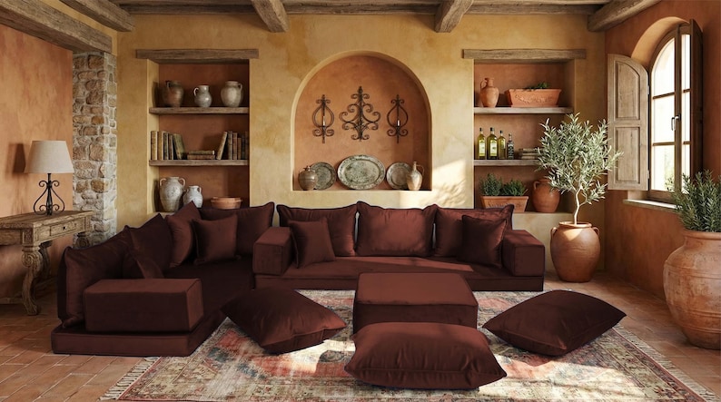 May include: A living room scene featuring a modular sectional sofa in a deep burgundy color. The room has a rustic, Mediterranean style with terracotta walls, wooden beams, and decorative shelves filled with pottery and books. The sofa is accompanied by matching ottomans and throw pillows.