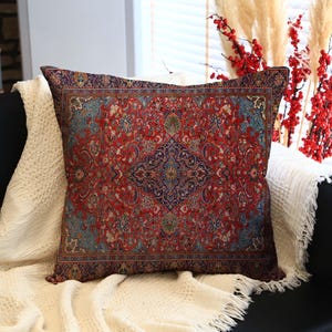Pode incluir: Uma almofada retangular com um padrão detalhado e ornamentado em tons de vermelho, azul e castanho. A almofada repousa sobre um cobertor branco texturizado, colocado num sofá preto. Ramos decorativos com bagas vermelhas no fundo.