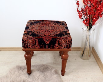 May include: A small, square ottoman with a dark, ornate patterned top in shades of black and rust. The wooden legs are a warm brown color. The ottoman sits on a light-colored floor, with a fluffy rug in the foreground.