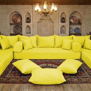 May include: A large, modular yellow sofa with numerous cushions and a low profile. The sofa is arranged in a U-shape, with additional cushions on the floor. The room features a patterned rug, wooden doors, and decorative wall niches.