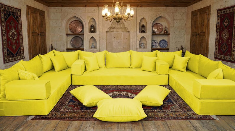 May include: A large, modular yellow sofa with numerous cushions and a low profile. The sofa is arranged in a U-shape, with additional cushions on the floor. The room features a patterned rug, wooden doors, and decorative wall niches.