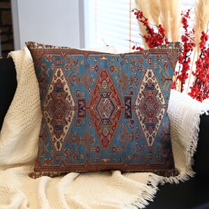 May include: A square throw pillow with a detailed, ornate pattern in shades of blue, red, and brown. The pillow is placed on a white blanket, resting on a black couch. Decorative elements include red berries and tall, dried grasses.