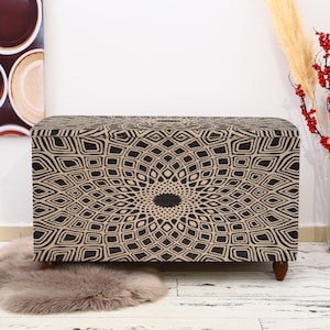 May include: A black and beige storage ottoman with a geometric pattern. The ottoman has a square shape and is sitting on a white wooden floor.