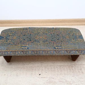 Könnte beinhalten: Ein niedriger, rechteckiger Fußhocker mit einer gemusterten blau-beige Stoffoberfläche. Der Stoff weist ein detailliertes, verziertes Design auf. Der Hocker wird von vier dunklen Holzbeinen getragen. Rechts befindet sich eine kleine Topfpflanze.