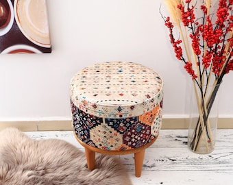 May include: A round, colourful patterned ottoman with wooden legs. The ottoman has a cream-coloured top with a geometric design and a multi-coloured bottom with a more intricate pattern. The ottoman is sitting on a white wooden floor.