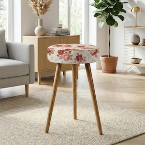 Puede incluir: Un taburete redondo con un asiento tapizado de color crema con un estampado floral en tonos rojos y rosas. El taburete se apoya sobre tres patas de madera delgadas y cónicas con un acabado marrón natural, proporcionando una base estable.