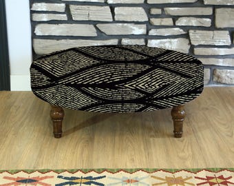 May include: An oval-shaped ottoman featuring a black and cream geometric design. It stands on four turned wooden legs, finished in a dark brown hue. The ottoman is placed on a light wood floor, with a patterned rug visible.