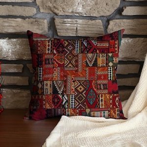 May include: Square decorative pillow with a patchwork design in shades of red, orange, and brown. The pillow features geometric and tribal patterns. The pillow is placed on a wooden surface with a stone background.