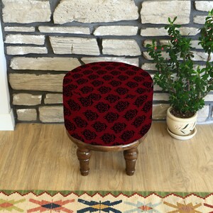 Puede incluir: Un pequeño taburete redondo de madera con una parte superior de tela burdeos con un estampado floral negro. El taburete tiene cuatro patas torneadas y está colocado sobre un suelo de madera. Una planta en maceta está a la derecha.
