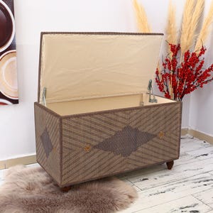 May include: A brown wicker storage chest with a diamond pattern and a beige fabric lid. The lid is open and the chest is sitting on a white wooden floor.