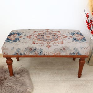 May include: A rectangular ottoman with a patterned fabric top in shades of blue, orange, and cream. The wooden frame and legs are a warm brown color. The ottoman is placed on a light-colored floor with a faux fur rug.
