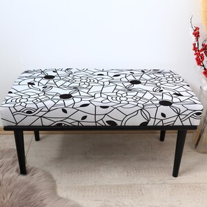 May include: A rectangular bench with a black metal frame and legs. The bench seat has a white fabric with a black geometric pattern. The design includes circular and linear shapes. The bench is a decorative furniture piece.