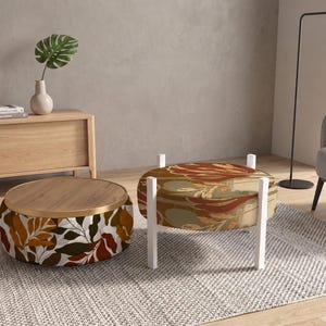 May include: Two round ottomans with patterned fabric. One ottoman features a white base with brown and dark green leaf prints and a wooden top. The other ottoman has a brown and green floral pattern and is supported by four white legs.