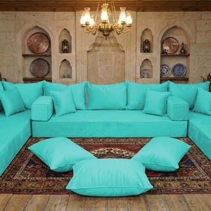 Peut inclure: Un grand canapé d'angle turquoise avec de nombreux coussins et oreillers. Le canapé est disposé en forme de L et repose sur un tapis à motifs. La pièce a un design rustique avec des planchers en bois et des murs en pierre.