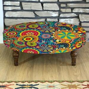 May include: An oval ottoman with a vibrant, colorful floral pattern in shades of blue, yellow, red, and green. The ottoman has dark brown wooden legs and sits on a light-colored wooden floor. The background features a stone wall.