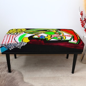 May include: A black bench with a colorful abstract design on the top. The artwork features a mix of red, green, yellow, and blue shapes. The bench has four black legs and is placed on a light-colored wooden floor.