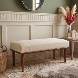 May include: A cream-coloured bench with a vertically striped pattern and dark brown wooden legs. A woven basket with leather handles sits to the left, and a small side table with a vase of flowers is on the right.