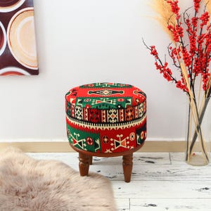 May include: A round ottoman with a colorful patterned fabric top in red, green, and black. The ottoman has a wooden base with four short, turned legs. The fabric design features geometric shapes and patterns. The ottoman is a decorative furniture piece.