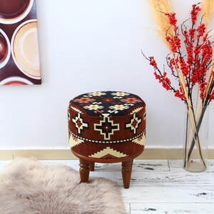 Può includere: Un pouf rotondo imbottito con un motivo geometrico nei toni del marrone, nero e crema. Il pouf ha una base in legno con quattro gambe tornite. Rami decorativi con bacche rosse in un vaso trasparente.