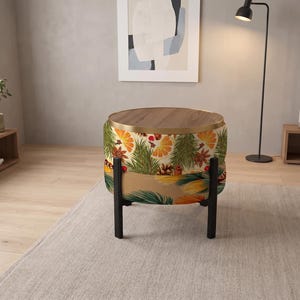 May include: A round ottoman with a wooden top and a gold metal rim. The base is upholstered in a fabric featuring a pattern of orange slices, pine branches, pinecones, and star anise. It stands on four black legs in a room setting.
