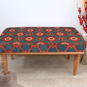 K&ouml;nnte beinhalten: Eine Holzbank mit einem blauen und roten gemusterten Kissen. Das Kissen hat ein geometrisches Muster mit floralen Motiven. Die Bank hat vier Beine und ist aus Naturholz.