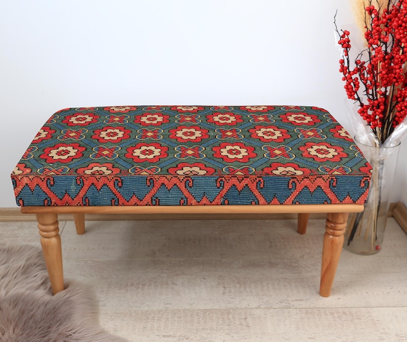 K&ouml;nnte beinhalten: Eine Holzbank mit einem blauen und roten gemusterten Kissen. Das Kissen hat ein geometrisches Muster mit floralen Motiven. Die Bank hat vier Beine und ist aus Naturholz.