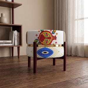 May include: A round ottoman with a white base featuring a colorful pomegranate illustration on the top section and a blue and gold eye motif on the bottom section, supported by four dark brown wooden legs with a gold metal band in the center.