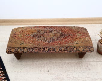 May include: A rectangular, low wooden footstool with a patterned rug top. The rug features a central medallion design in shades of red, brown, and navy blue. The stool has four short, dark wooden legs. A small potted plant is to the right.