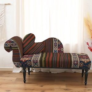 May include: A decorative chaise lounge with a patterned fabric in shades of red, brown, and blue. The lounge has a curved back and armrest, and sits on black legs. A macrame wall hanging and a small side table are in the background.