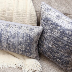 Könnte beinhalten: Zwei dekorative Kissen mit einem verblassten blauen und weißen Muster. Die Kissen liegen auf einem hellbraunen Sofa mit einer weißen Wolldecke.