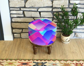 Puede incluir: Un pequeño taburete redondo de madera con una parte superior estampada con motivos geométricos coloridos. El diseño presenta formas superpuestas en tonos de rosa, azul y morado. El taburete tiene cuatro patas torneadas de color marrón oscuro. Una pequeña planta en maceta está cerca.