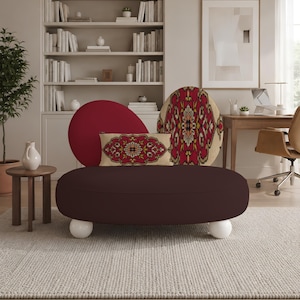 May include: A round, dark brown upholstered sofa featuring two white spherical feet. The sofa is accented with a rectangular throw pillow and a circular cushion, both displaying a detailed red, beige, and black floral medallion pattern.