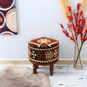 Può includere: Un pouf rotondo imbottito con un motivo geometrico marrone e crema. Il pouf ha una base in legno con quattro gambe corte e tornite. Bacche rosse decorative ed erba alta sono in un vaso trasparente sullo sfondo.