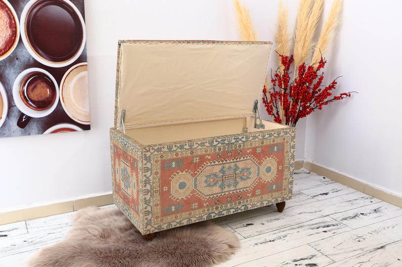 May include: A decorative storage ottoman with a hinged lid. The ottoman is covered in a colorful patterned fabric with a red, blue, and beige color scheme. The lid is open, revealing a beige fabric lining.